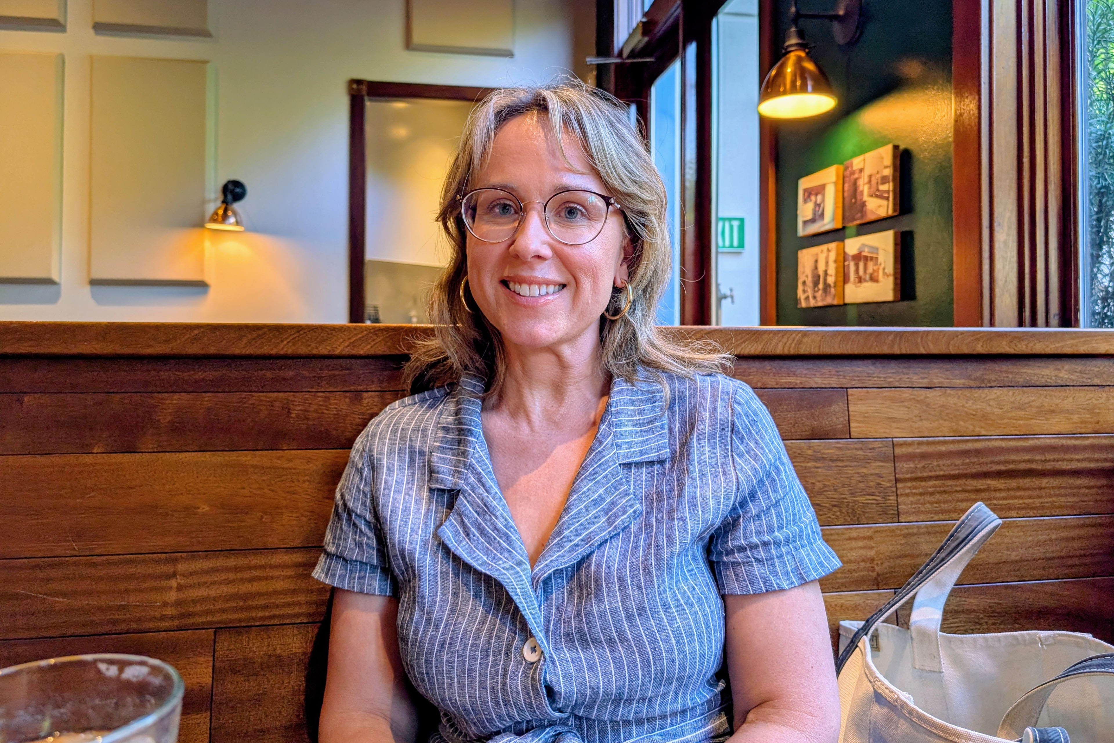 A photo of a woman wearing a blouse and smiling in the direction of the photographer, who she is sitting across from at a cafe.