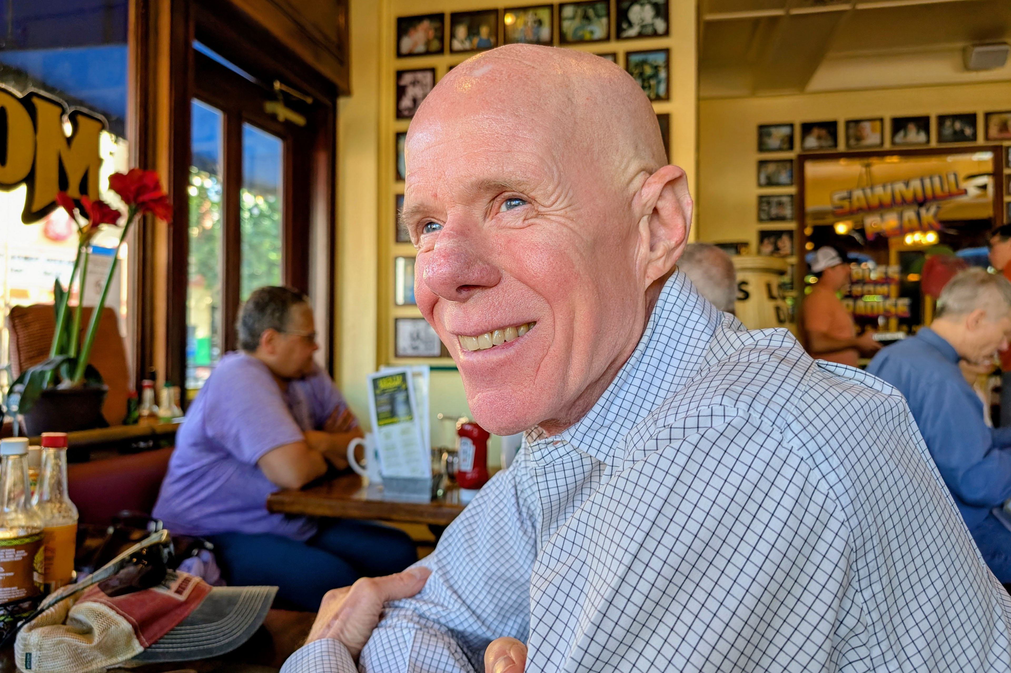 A photo of an older man wearing a striped button down. He is sitting at a cafe and smiling.