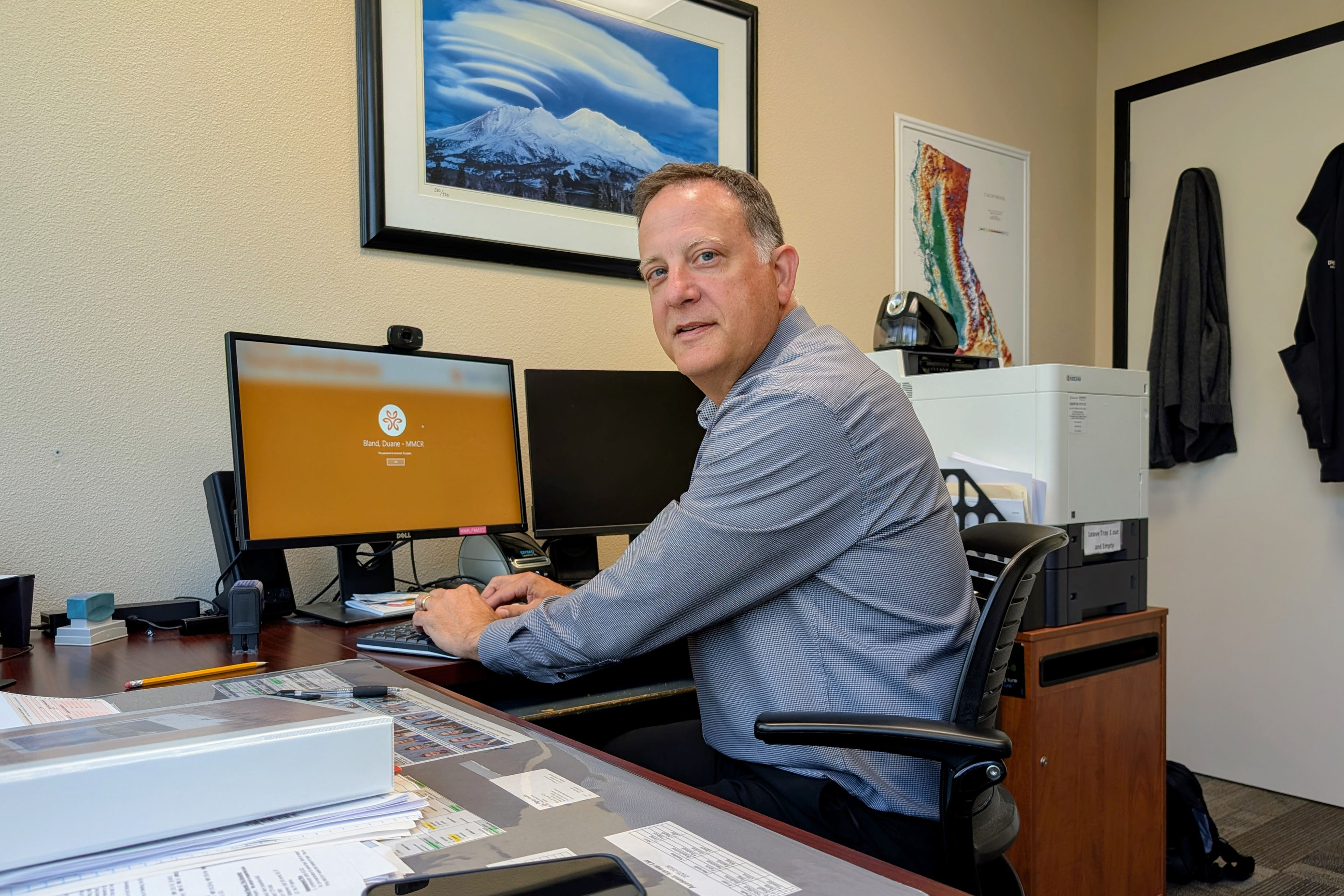 A photo of a man sitting at a desk in front of two computer screens. He turns to face the photographer with a gentle expression.