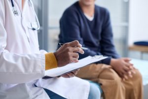 A photo of a doctor's appointment. A doctor reads off notes from a clipboard as a patient on the exam table listens.